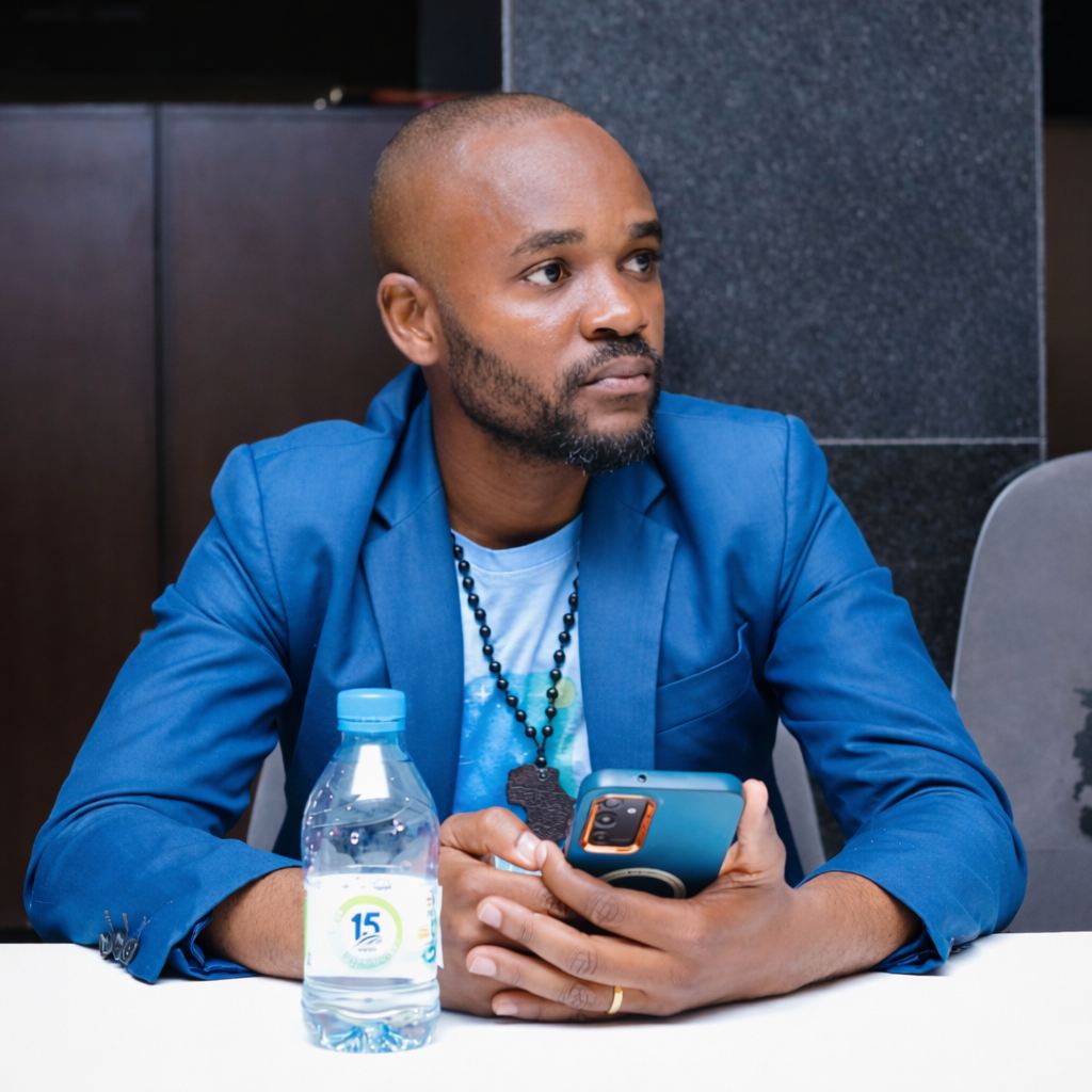 Man seated indoors holding a smartphone, wearing a blue jacket and necklace.