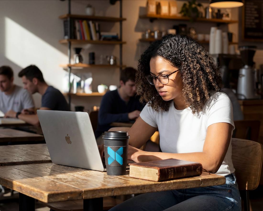 Student studying on a laptop with a Bible and an x3 Coffee cup at a café table.