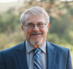 Smiling older man with gray hair and glasses wearing a blue suit and tie, photographed outdoors.
