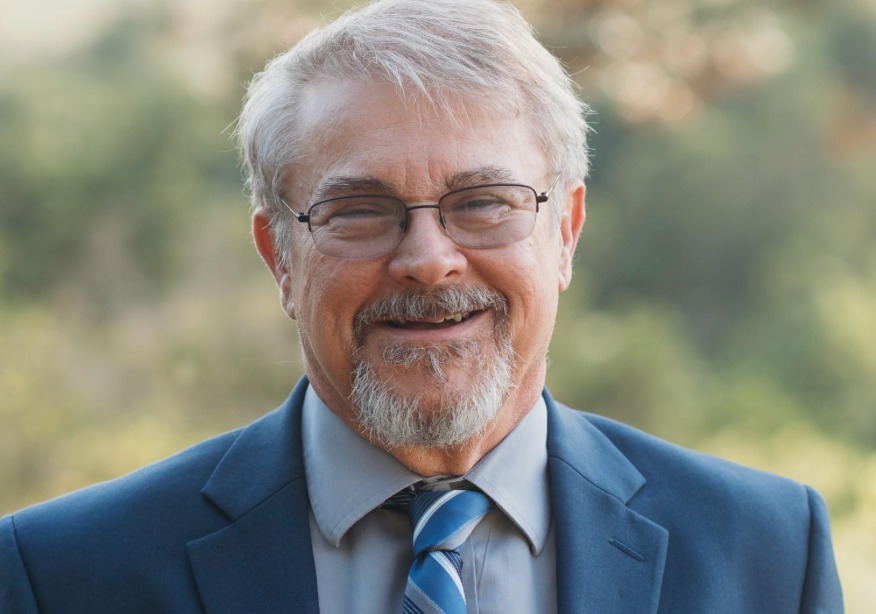 Person smiling outdoors wearing glasses and a suit jacket.