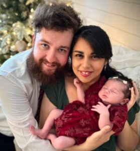 Smiling couple holding a newborn baby dressed in a red outfit, photographed indoors.