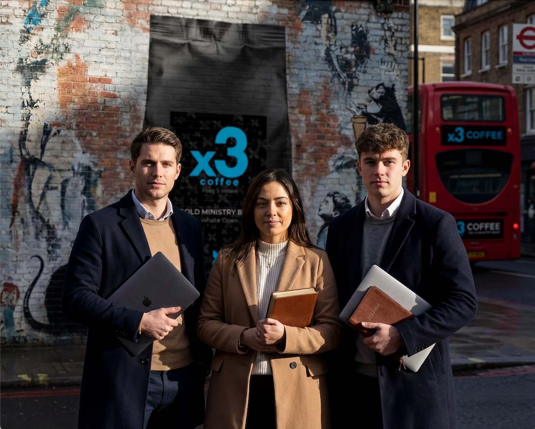 Three students holding books and laptops standing outdoors near an x3 Coffee sign.