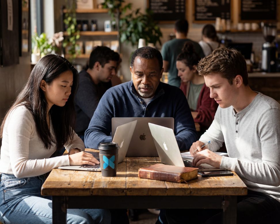 Small group of students studying together with laptops, a Bible, and an x3 Coffee cup at a café table.