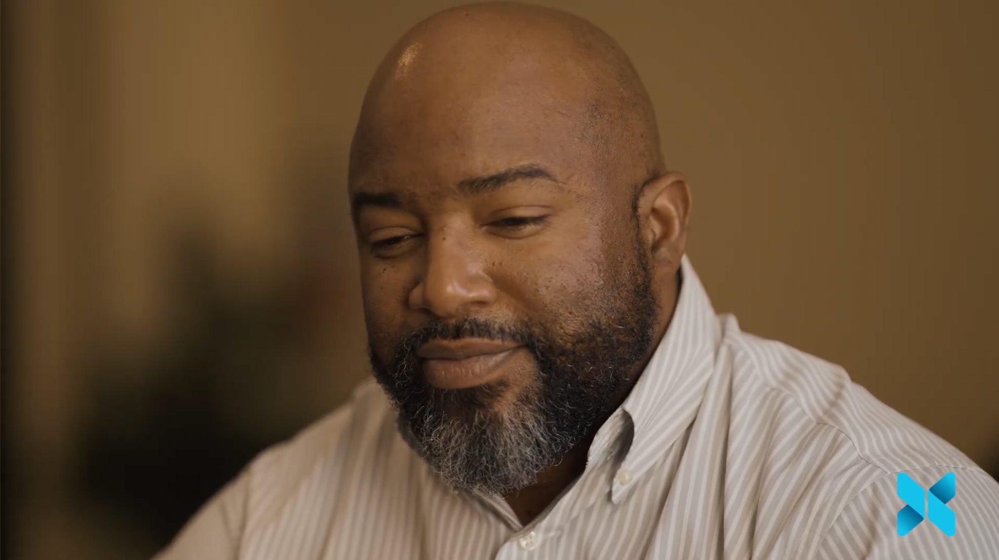 A man with a beard looks downward thoughtfully in a softly lit indoor setting.