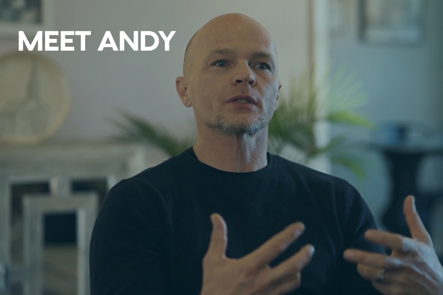Man speaking and gesturing with his hands in a softly lit indoor setting, wearing a dark shirt, with the text “MEET ANDY” displayed on the left side.