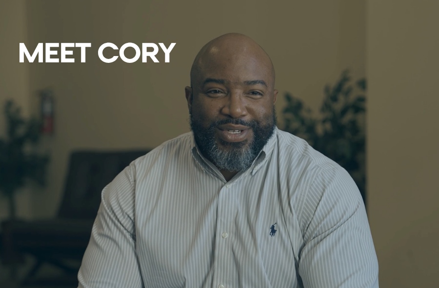 Man smiling in a warmly lit indoor setting, wearing a striped button-up shirt, with the text “MEET CORY” displayed on the left side.