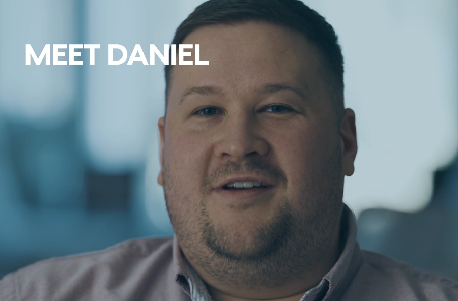 Man smiling in a softly lit indoor setting, wearing a collared shirt, with the text “MEET DANIEL” displayed on the left side.