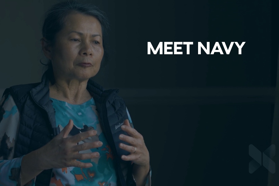 Woman speaking and gesturing with her hands in a softly lit indoor setting, wearing a patterned shirt and vest, with the text “MEET NAVY” displayed on the right side.