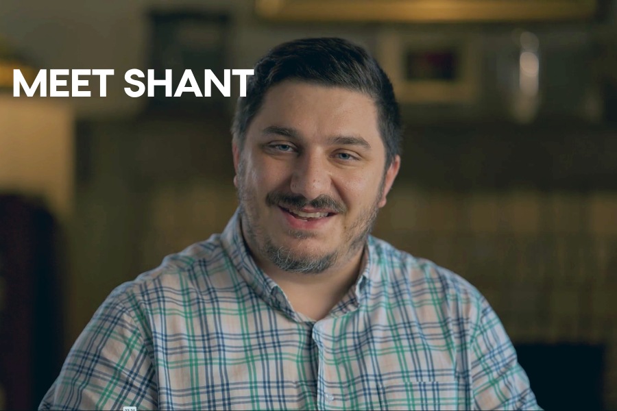 Man smiling in a warmly lit indoor setting, wearing a plaid button-up shirt, with the text “MEET SHANT” displayed on the left side.